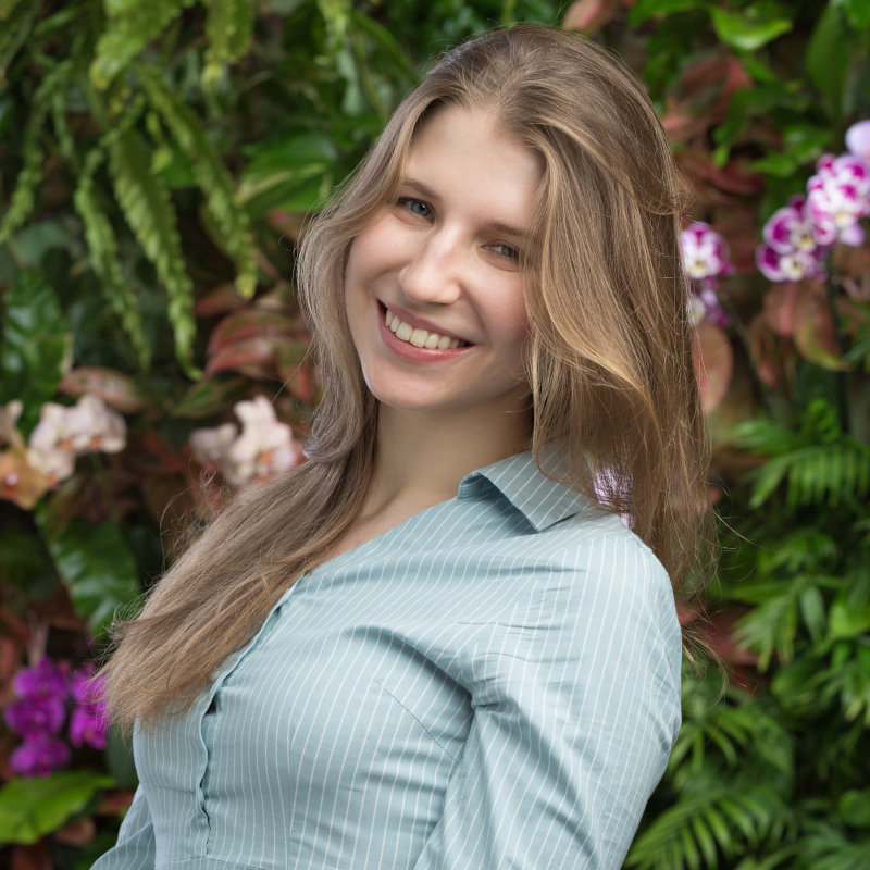 Sasha has long, blond hair, white skin and is wearing a blue shirt, with a floral background.