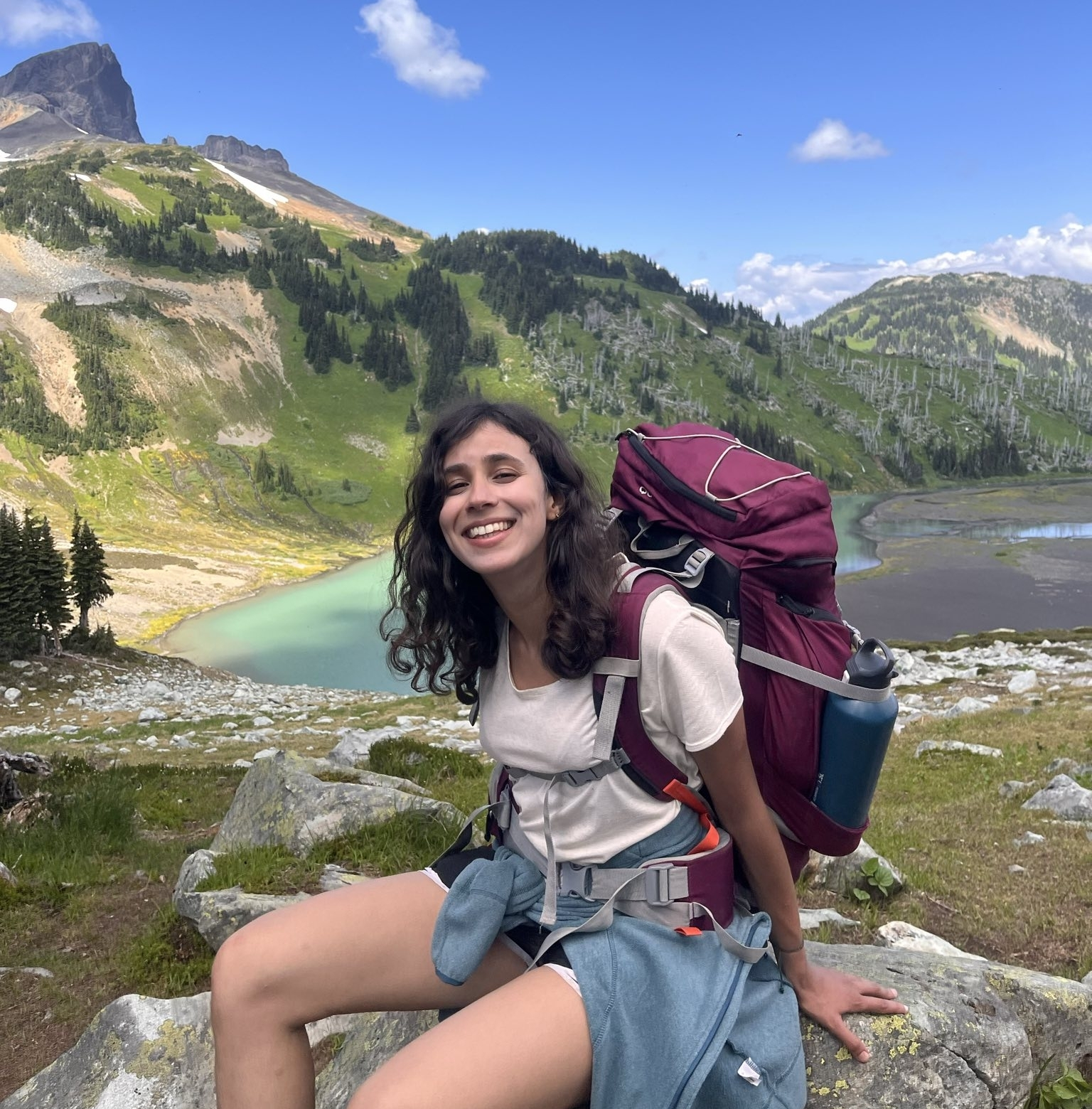 Léna has long, curly, dark hair, a big smile and is posing with a backpack in the mountains.